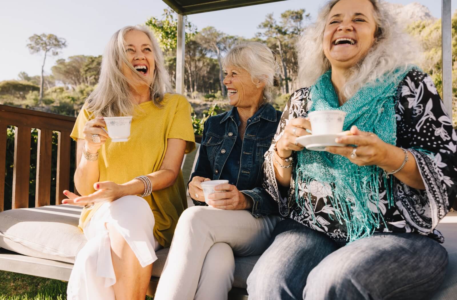 Three older adult women laugh together while enjoying cups of tea on an outdoor bench.