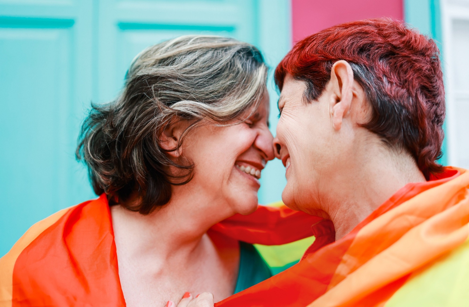 Two older adults embracing, wrapped in the LGBT flag.