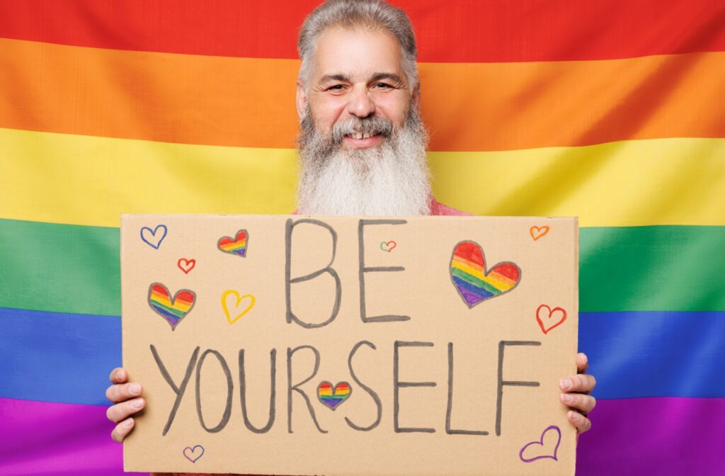 An older adult, standing in front of a rainbow flag, holds up a cardboard sign reading, be yourself
