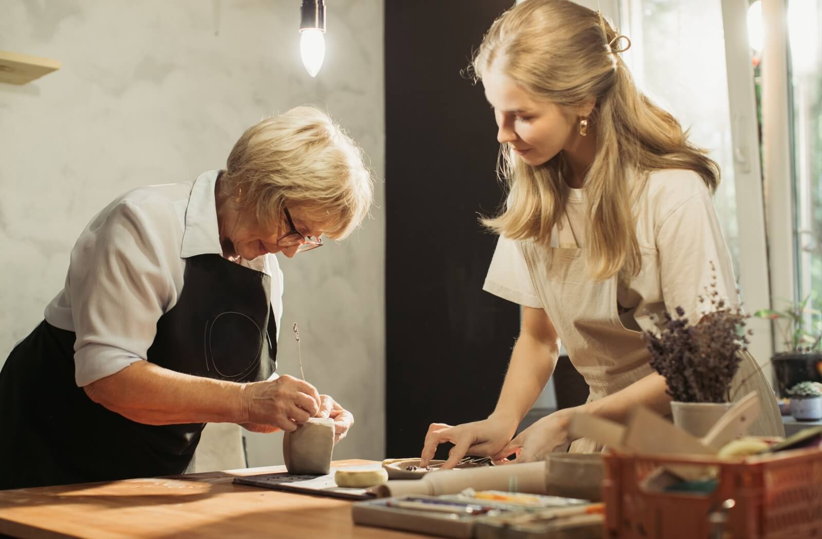 An older adult with dementia etches something into a wet clay sculpture while a caregiver smiles and watches