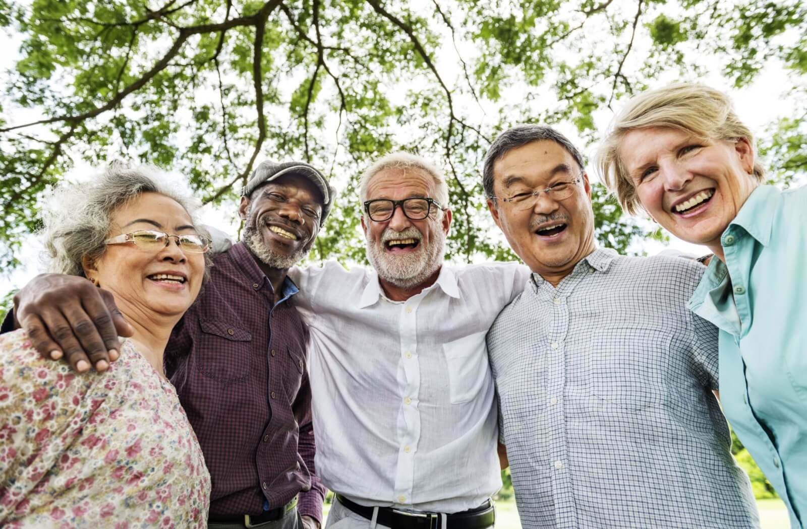 A group of older adults smiles in a semi-circle, taking a break in a social, outdoor walk to take a group picture.