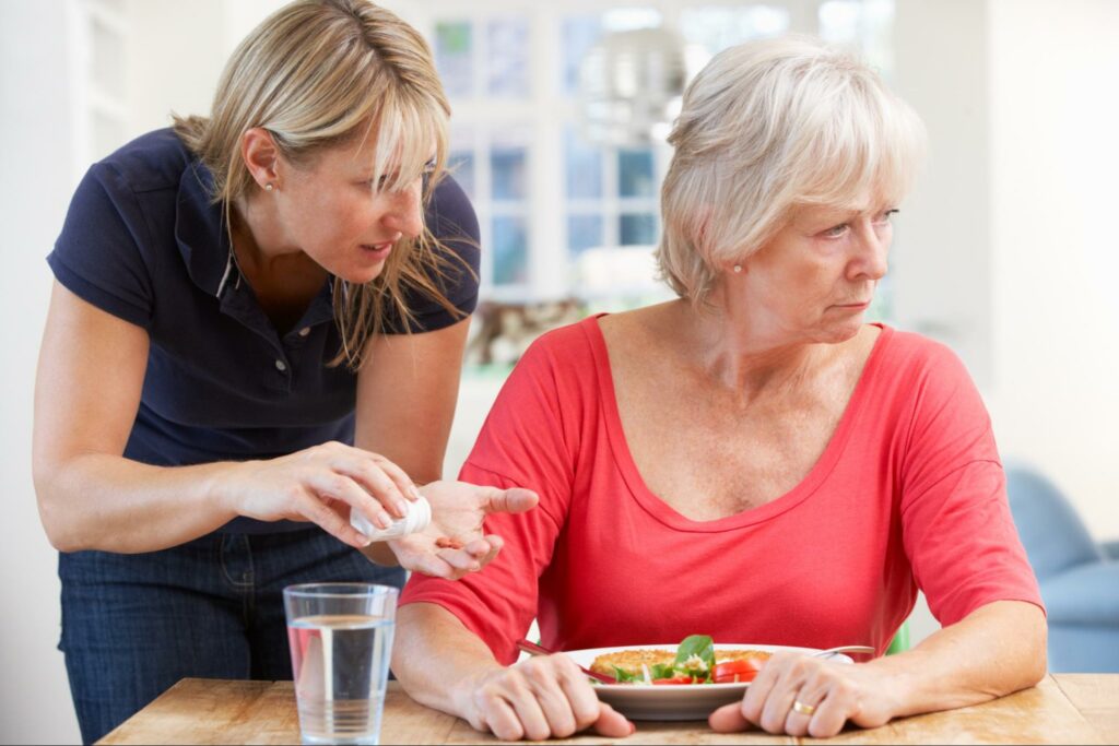 A senior women refusing her medication and ignoring her food, despite her insistent daughter.